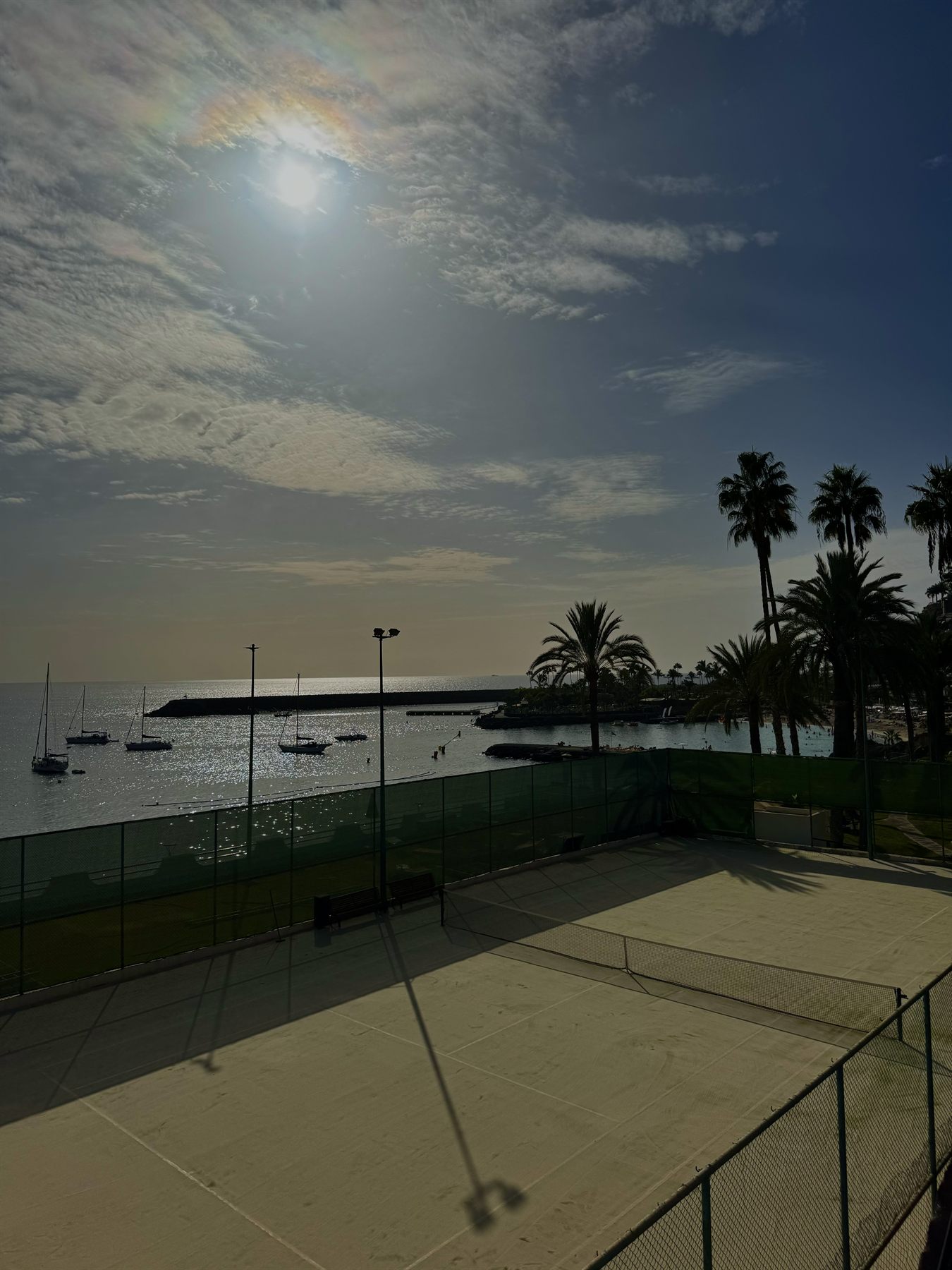 Tennisplatz der Anlage im Gegenlicht, dahinter die Bucht mit Segelbooten